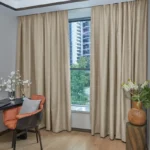 Neutral beige blackout curtains framing a city window next to a modern orange chair.